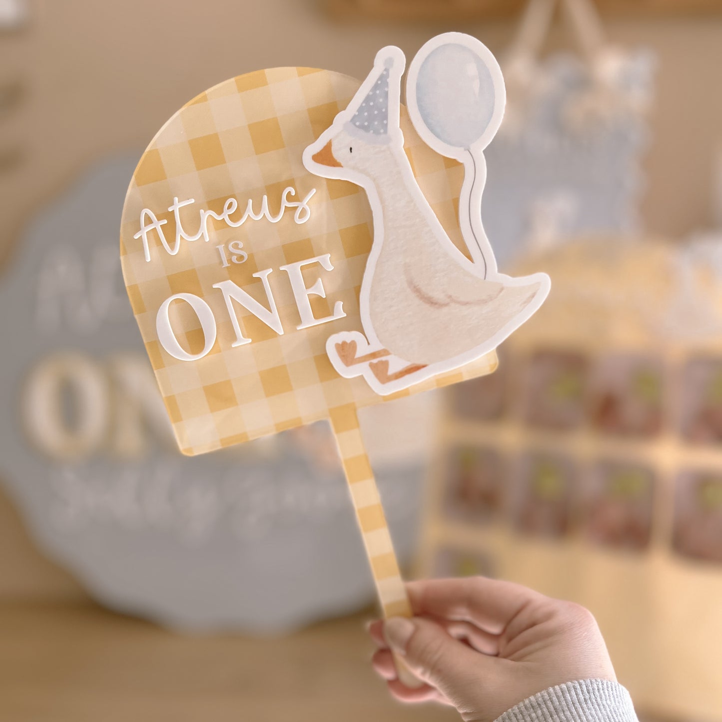 Hand holding a custom cake topper with a checkered design and text, blurred background
