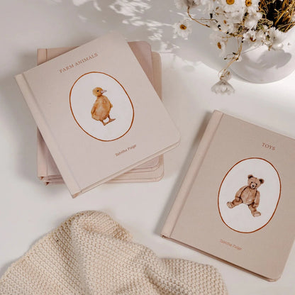 Two beige books with bear illustrations on a light surface with flowers and a blanket.
