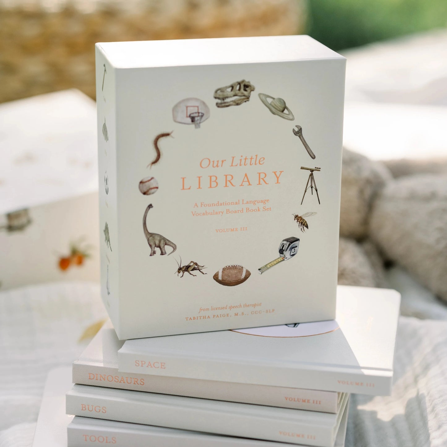 Stack of books titled 'Our Little Library' on a soft surface with a blurred natural background