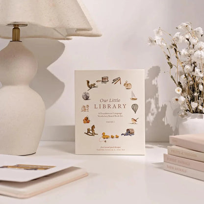 Book titled 'Our Little Library' on a table with a lamp and flowers in the background